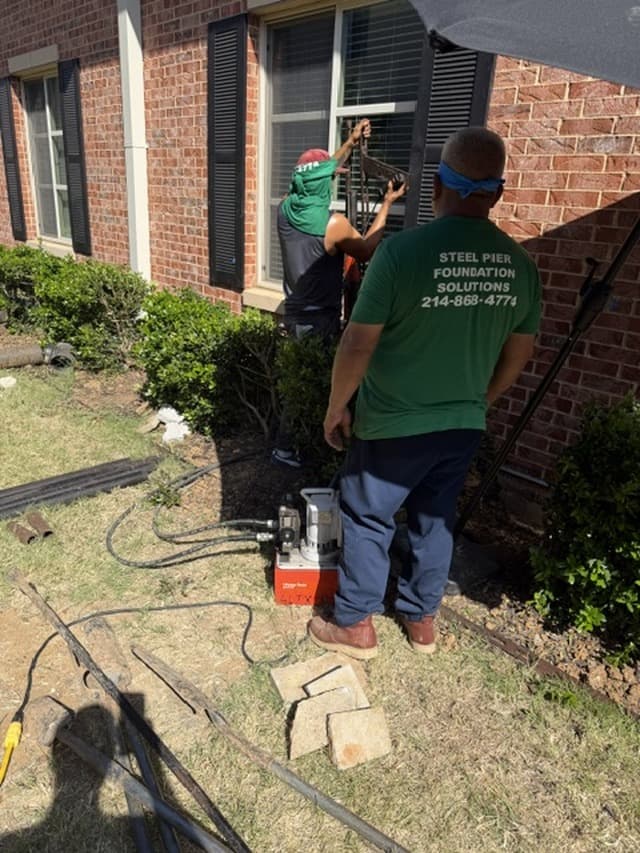 AlphaLift crew driving a steel pier with a hydraulic ram beside a brick home